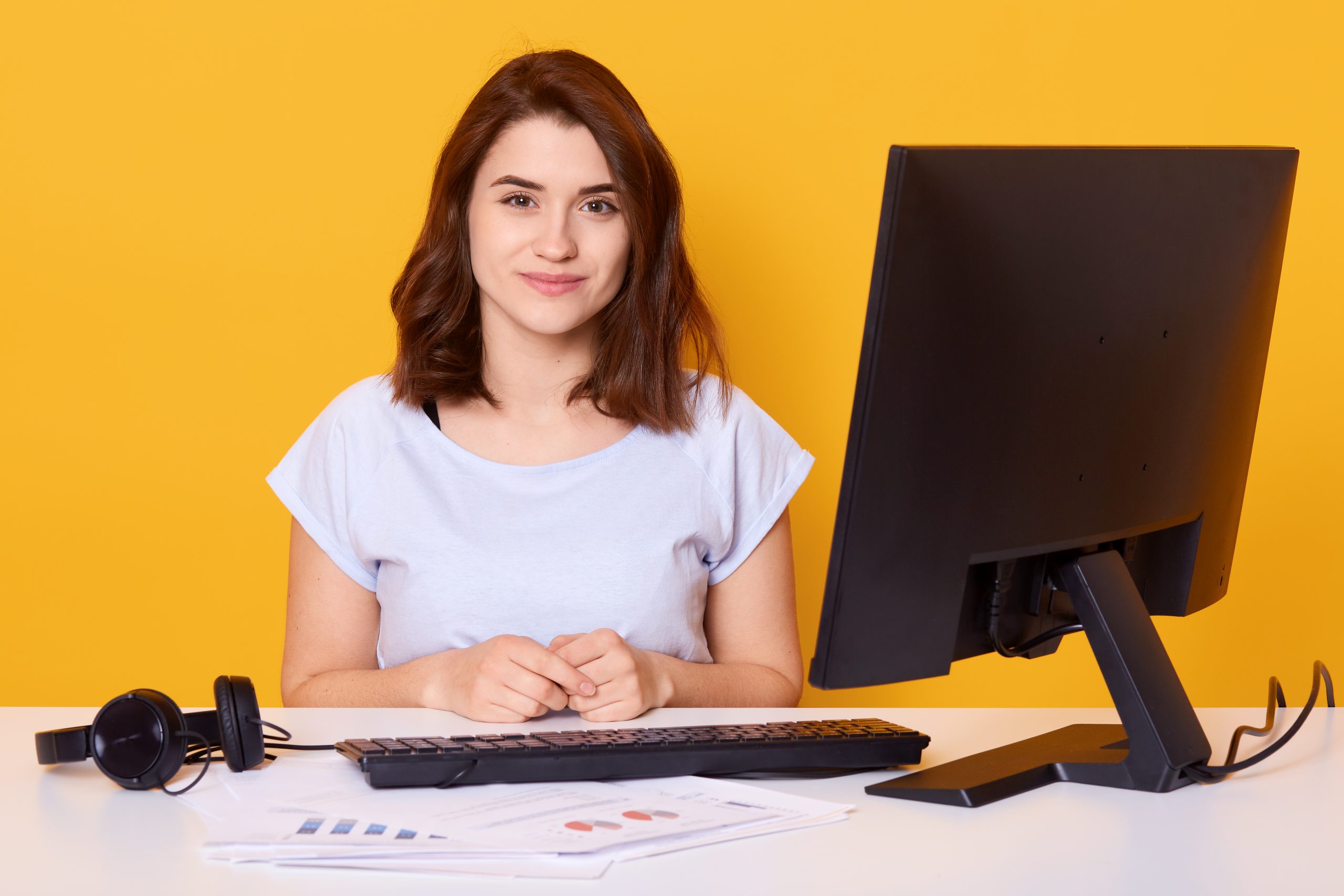 jovem posa sentada em sua mesa de trabalho, diante computador e planilhas impressas espalhadas pela mesa