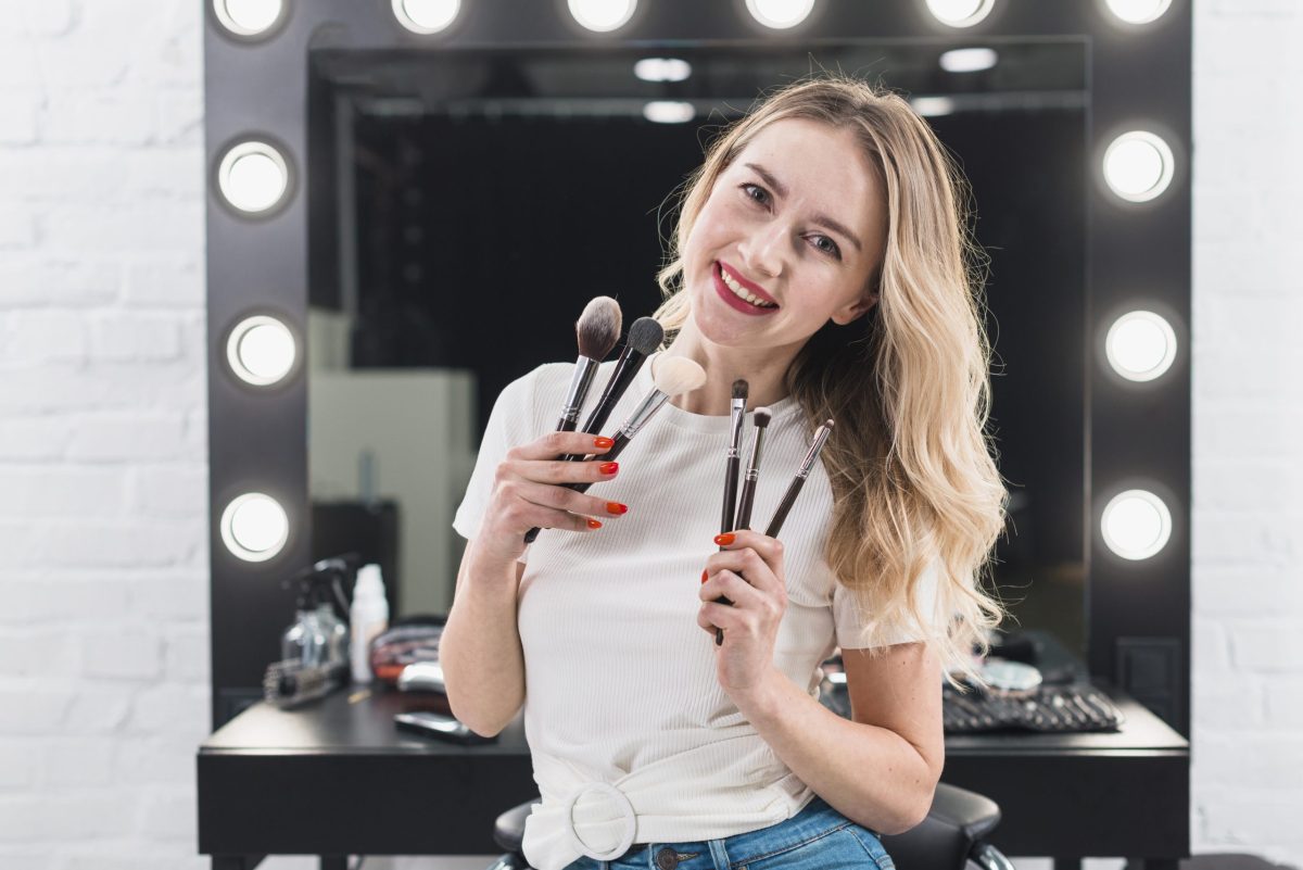 Maquiadora profissional posa em seu salão sorridente segurando alguns pinceis de maquiagem nas duas mãos