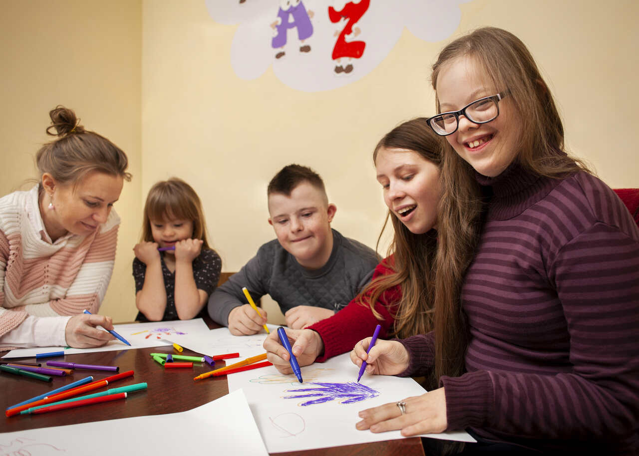 Menina com Sindrome de Down posa sorridente enquanto desenha com colegas na escola 
